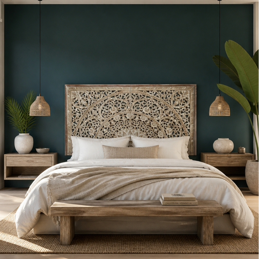 A styled bedroom featuring a bed with a highly detailed, ornate carved wooden headboard, dressed in white and beige bedding. The bed is set against a deep teal painted accent wall, flanked by matching wooden nightstands, woven pendant lights, and potted plants.